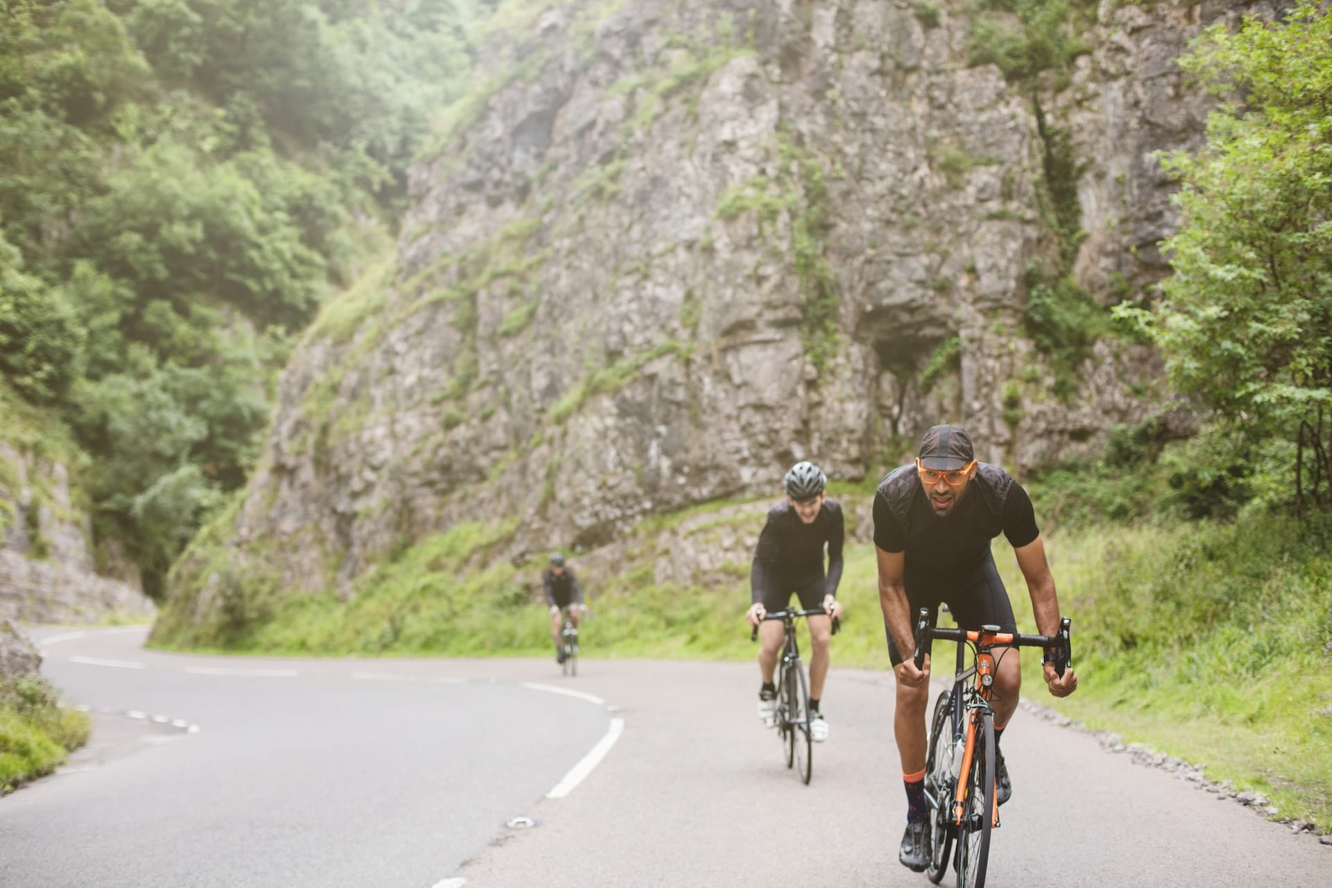 Cyclists riding road bikes on winding road through steep, green gorge with rock faces