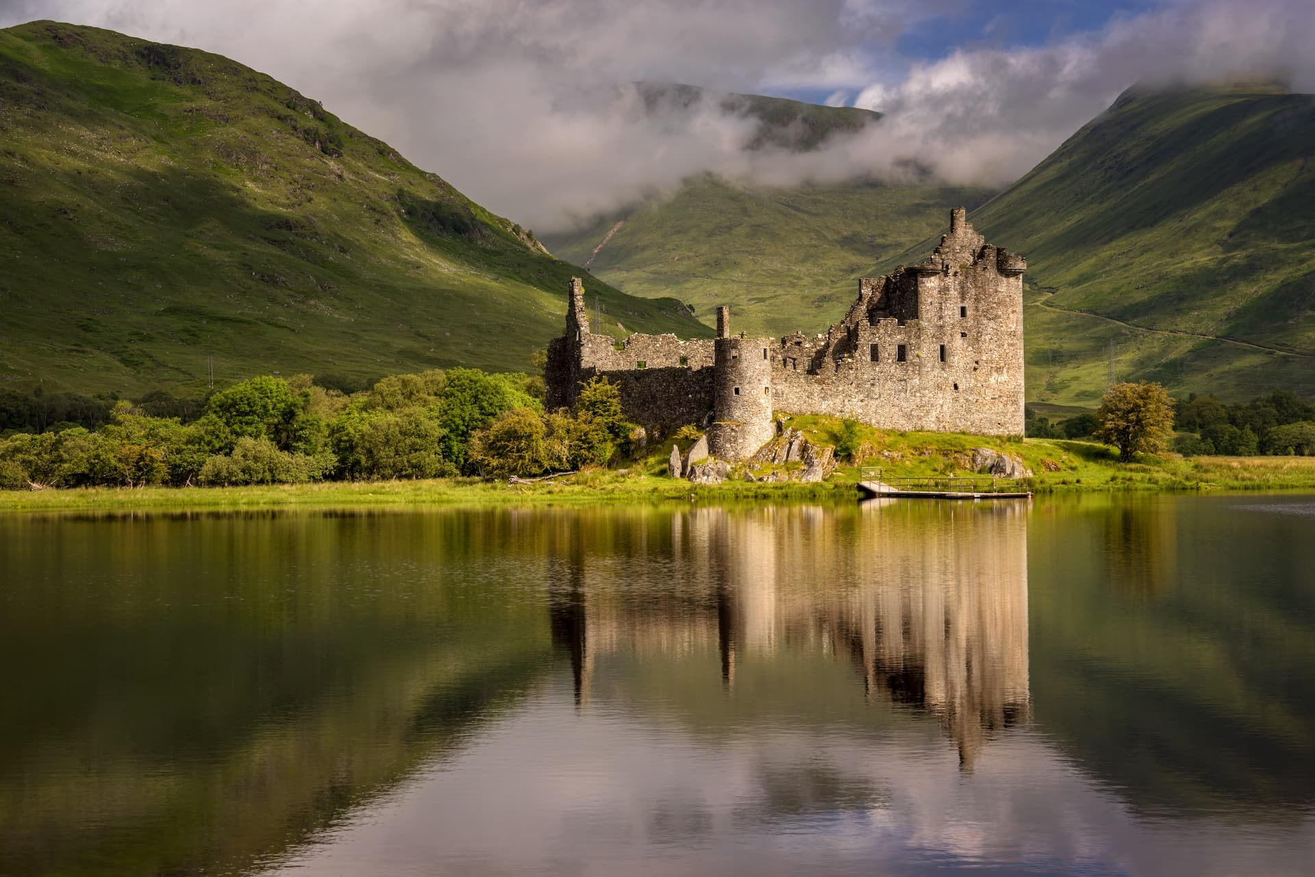 Kilchurn Castle ruins reflected in Loch Awe with green mountains shrouded in low clouds.