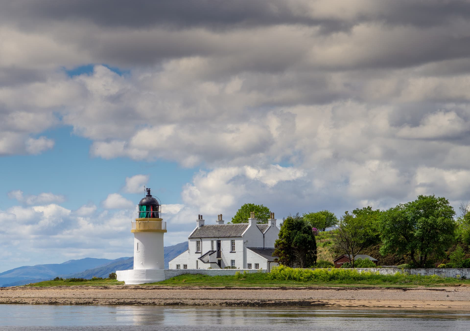 White lighthouse and keeper's cottage on a shore with mountains under dramatic clouds.