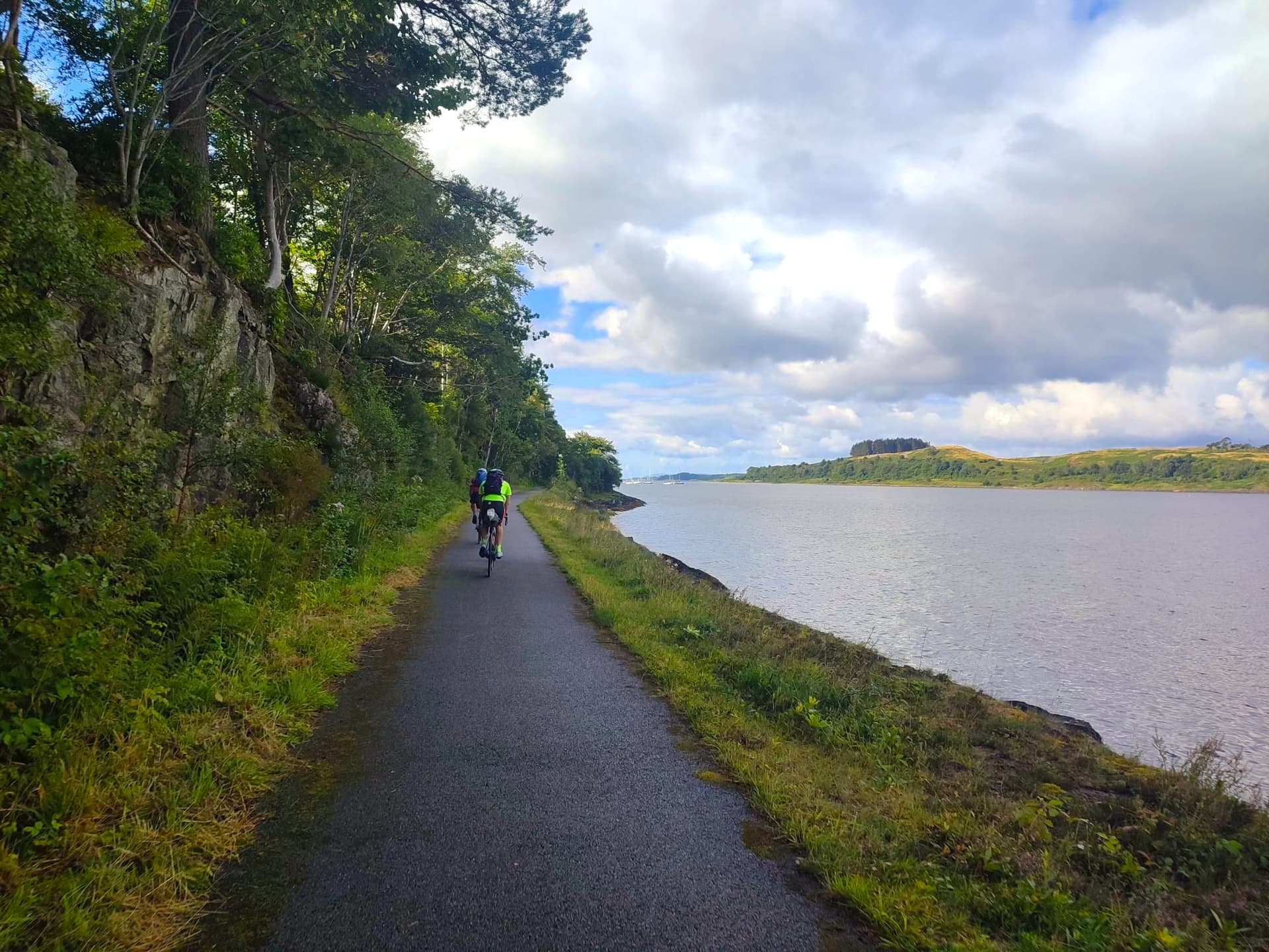 Cyclists on paved path beside water, with trees on a rocky bank and hills across the water.