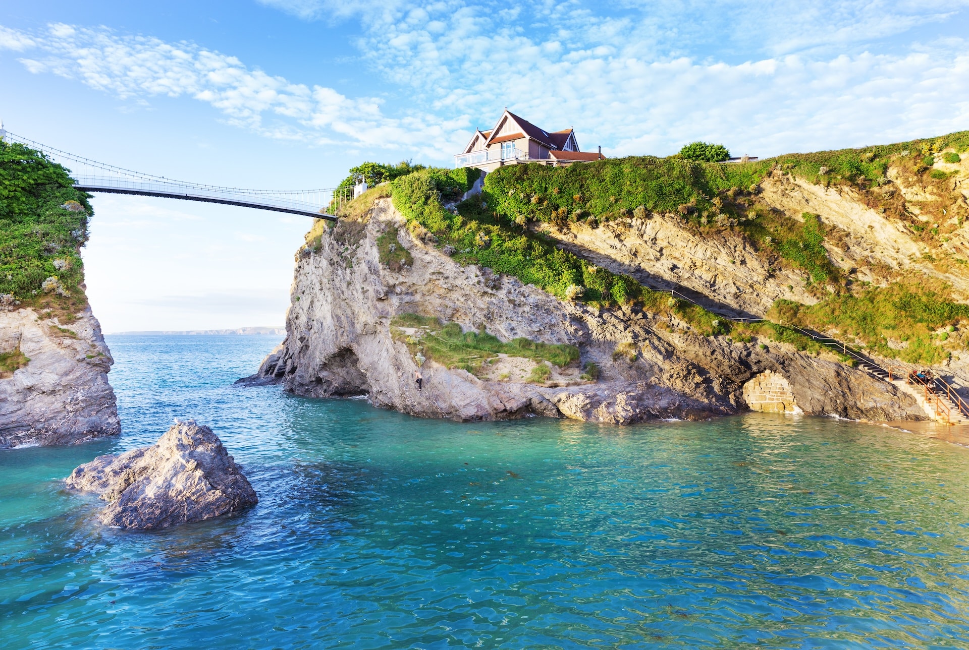 Suspension bridge connecting cliffs above turquoise sea with house on clifftop, Newquay.