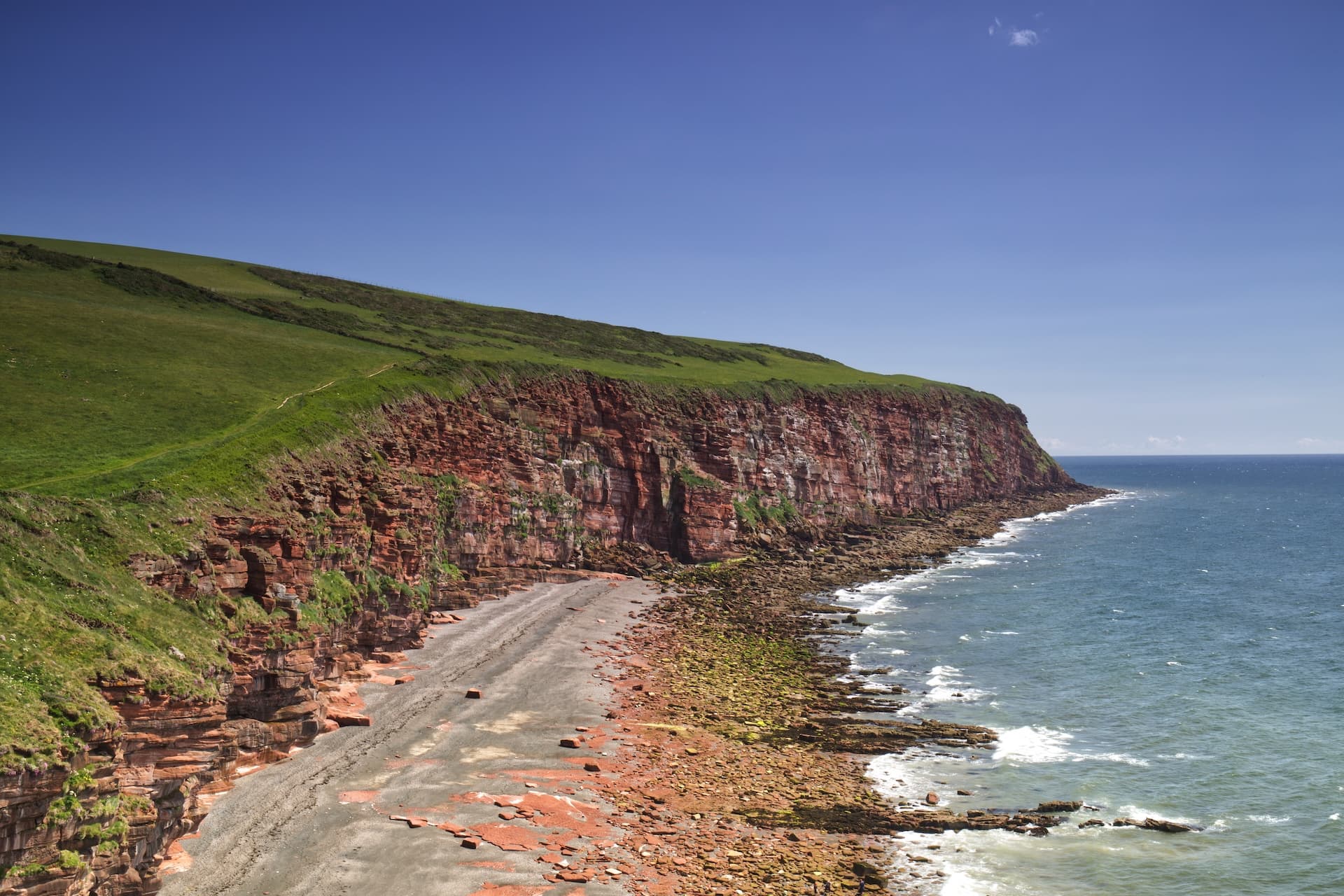 Red sandstone cliffs meeting the sea with a narrow rocky beach under a clear blue sky, Cumbrian Coast.
