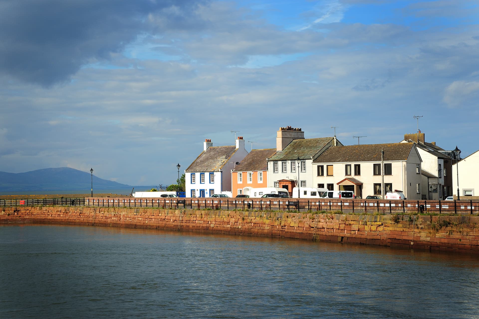 Colorful waterfront houses along a harbor wall with a distant mountain under a cloudy sky.