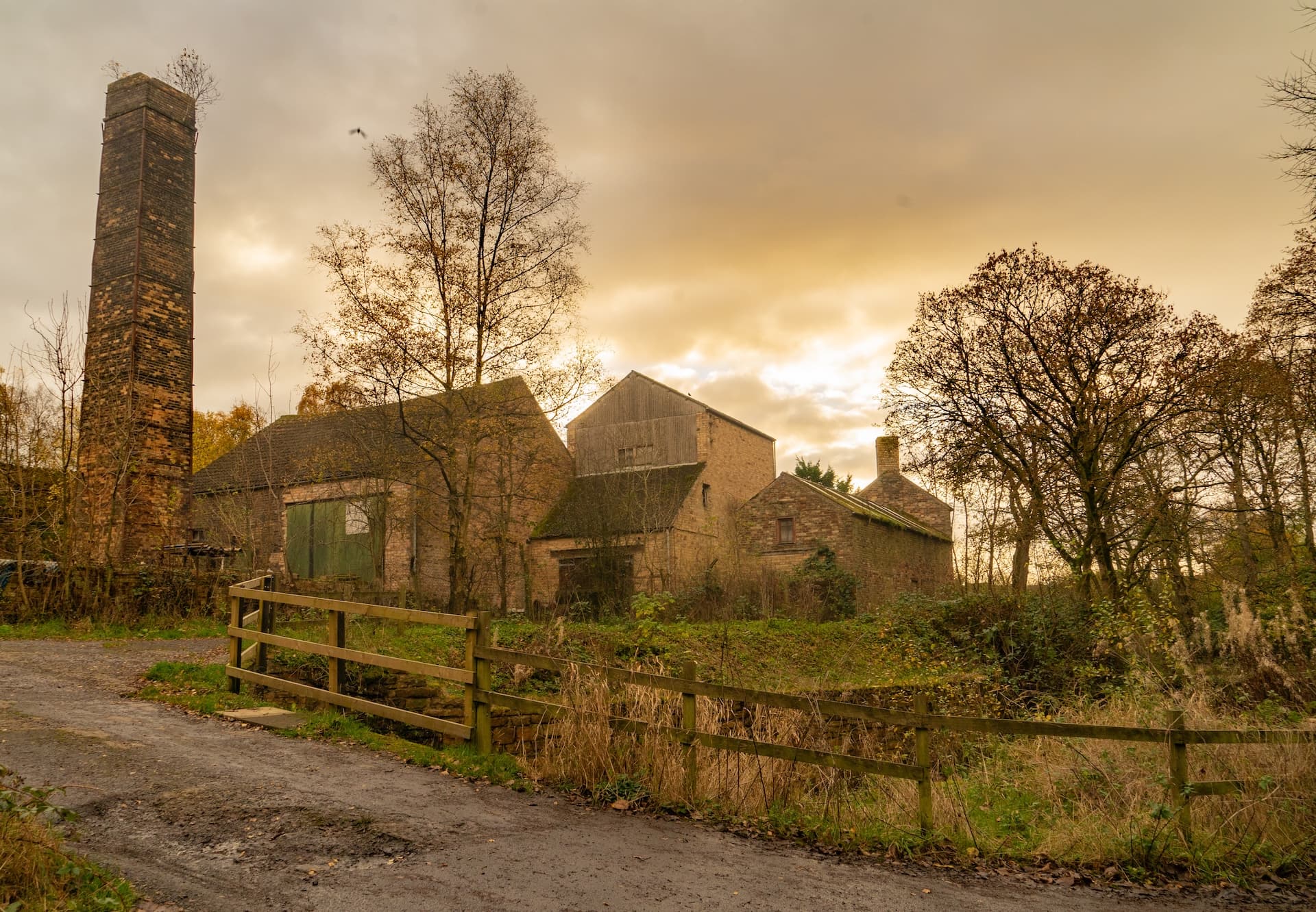 Brick industrial chimney and old farm buildings on a muddy track under an overcast sky.