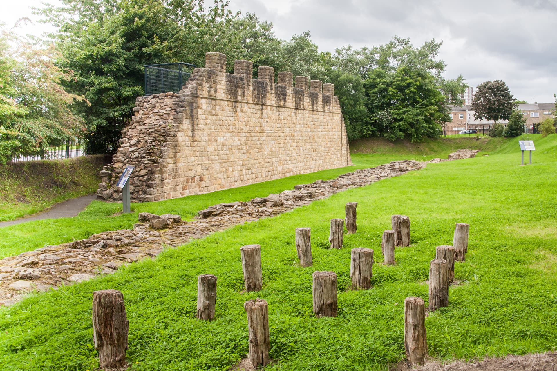 Remains of a stone wall with crenellations, wooden posts in grass, and informational signs at Wallsend Fort.
