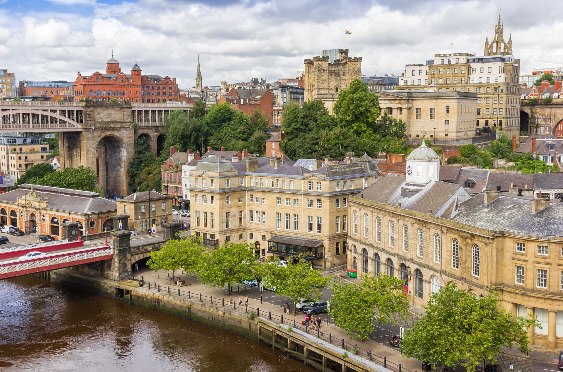 Cityscape of Newcastle featuring historic buildings, a castle keep, and a bridge over a brown river.