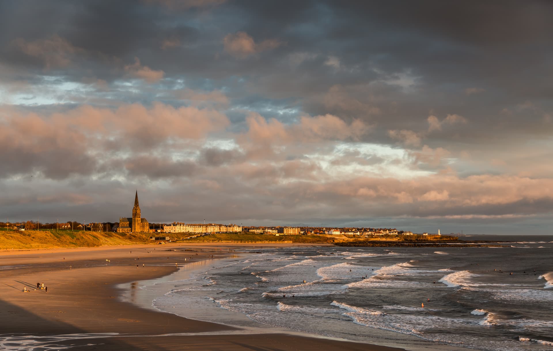 Surfers in ocean waves near Longsands Beach with town skyline under dramatic sunset clouds