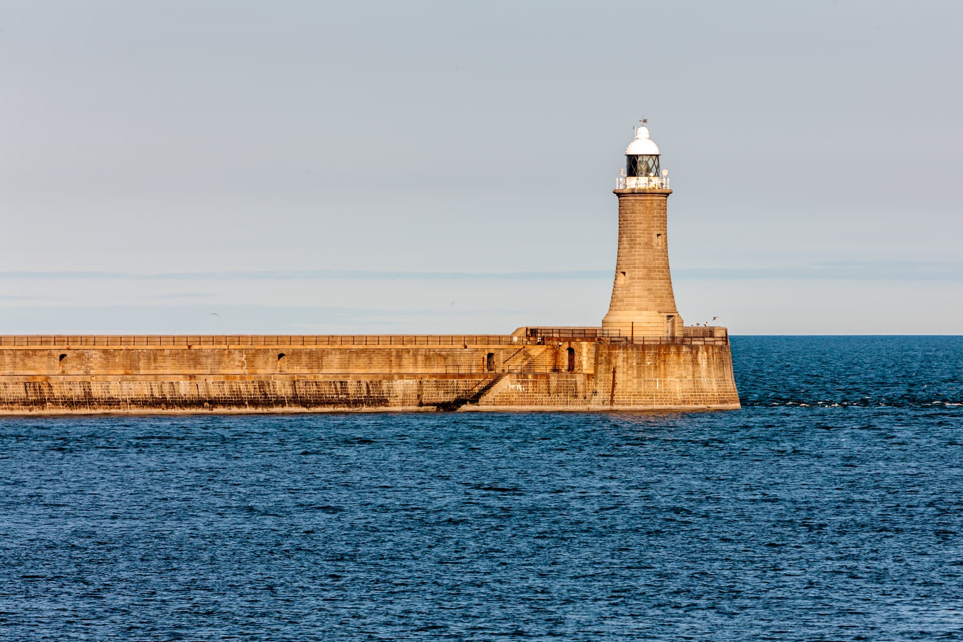 Stone lighthouse on a long sea pier against blue water and pale sky, Tynemouth