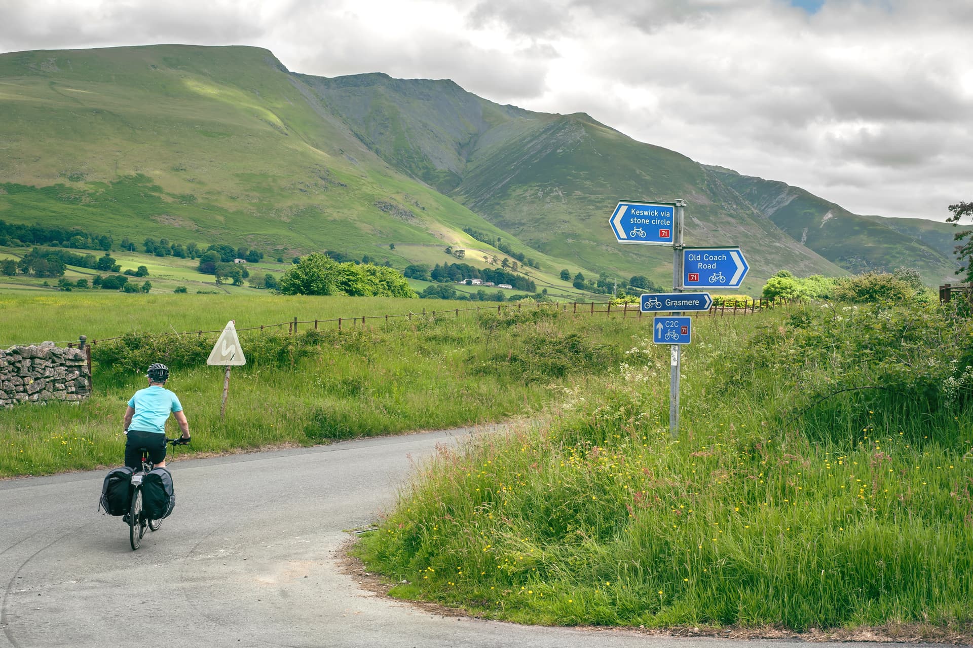 Cyclist with panniers riding past road signs toward Grasmere near green mountains.