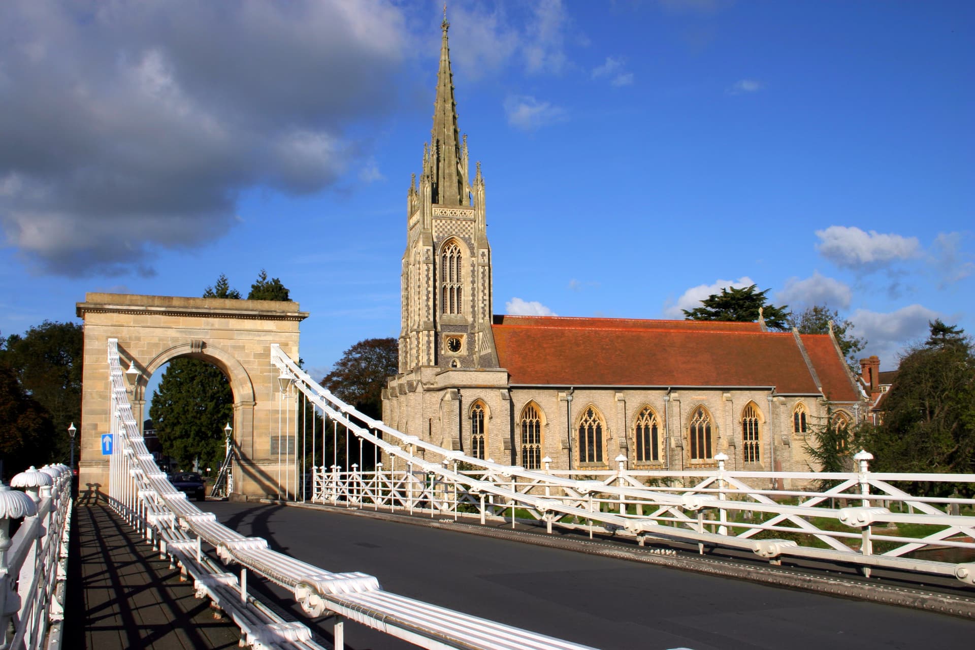 Suspension bridge with white railings leading toward a stone church with a tall spire.