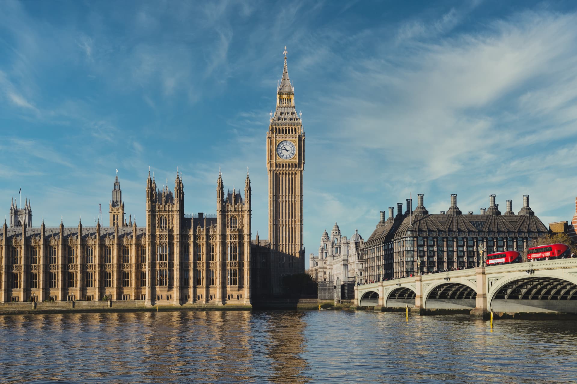 Big Ben clock tower and Palace of Westminster reflected in the River Thames with a red bus on the bridge.