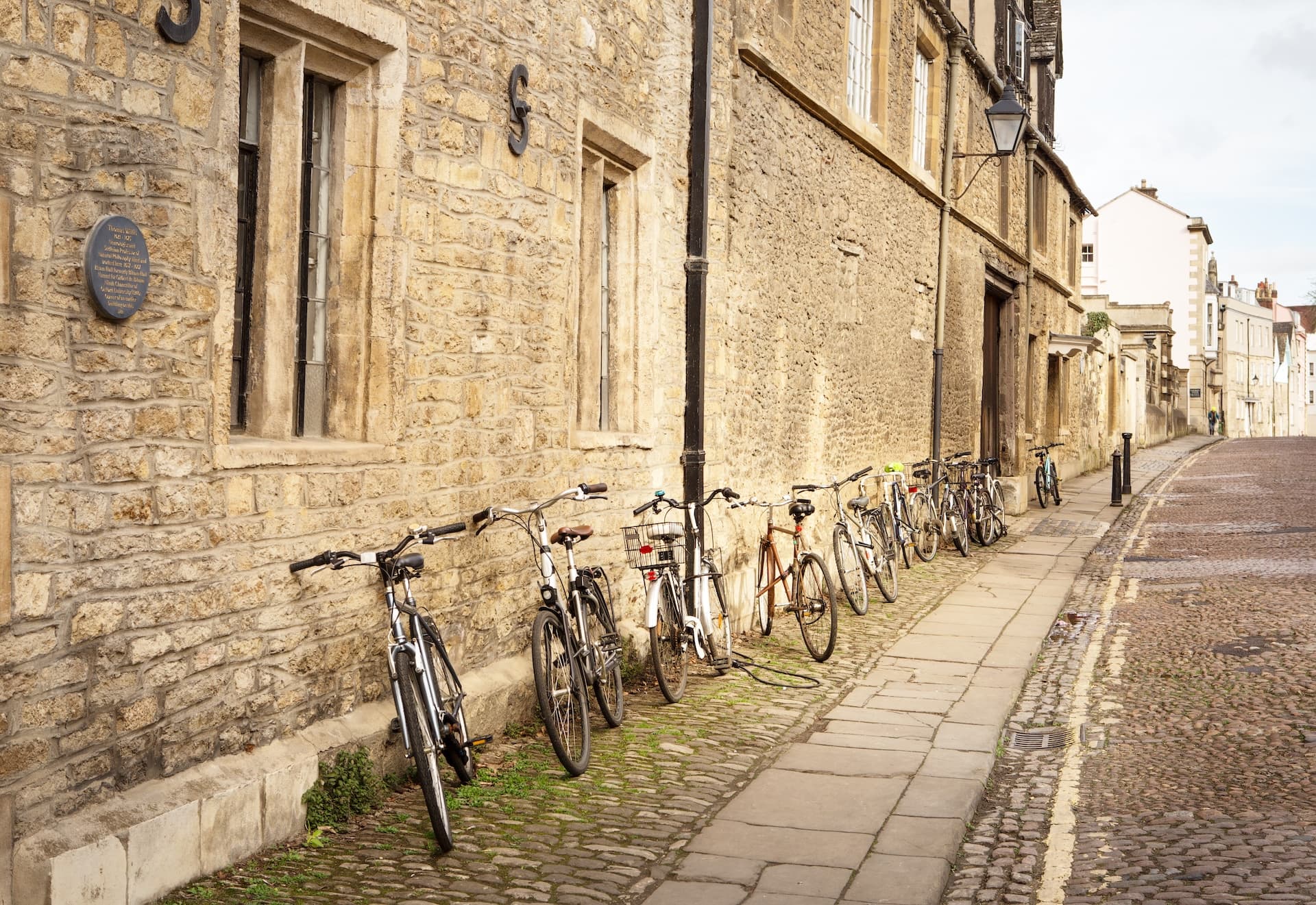 Bicycles parked along a stone wall on a cobblestone street in Oxford.
