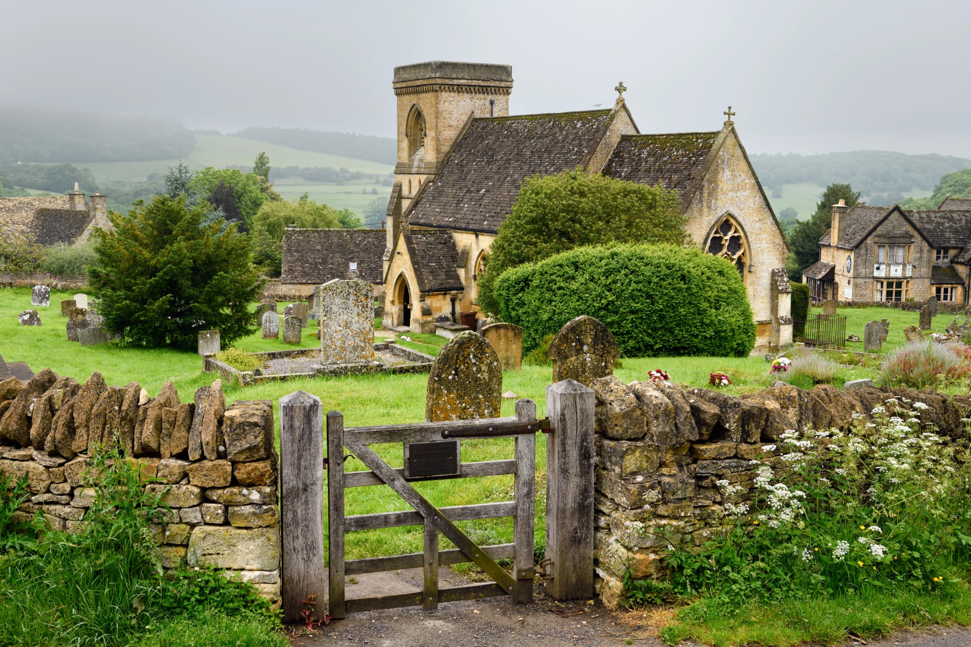 St Barnabas Anglican church with cemetery and stone wall gate in wet rainy weather in Snowshill Cotswold England
