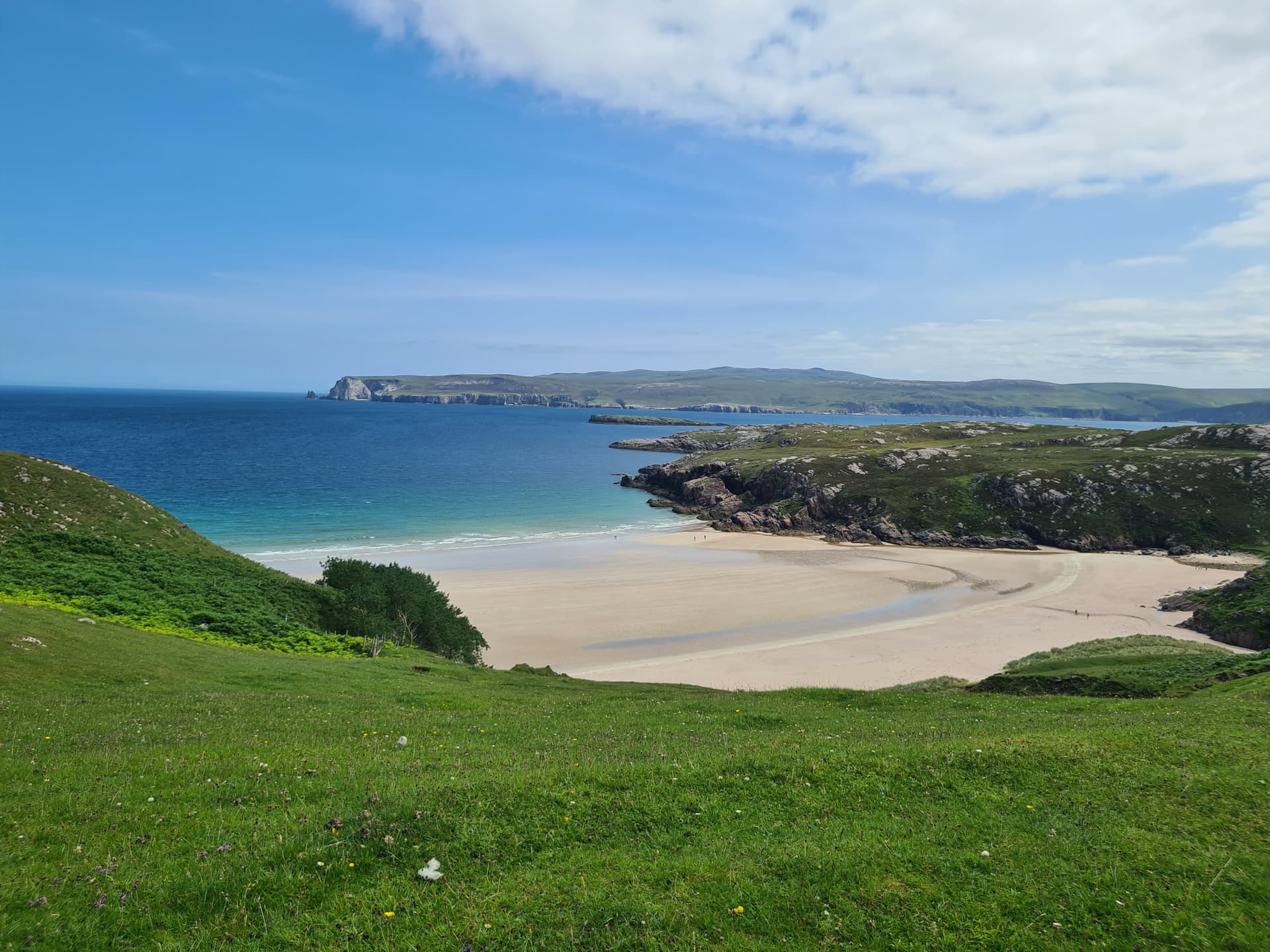 White sand bay and turquoise sea with green grassy hills and rocky coastline under a blue sky