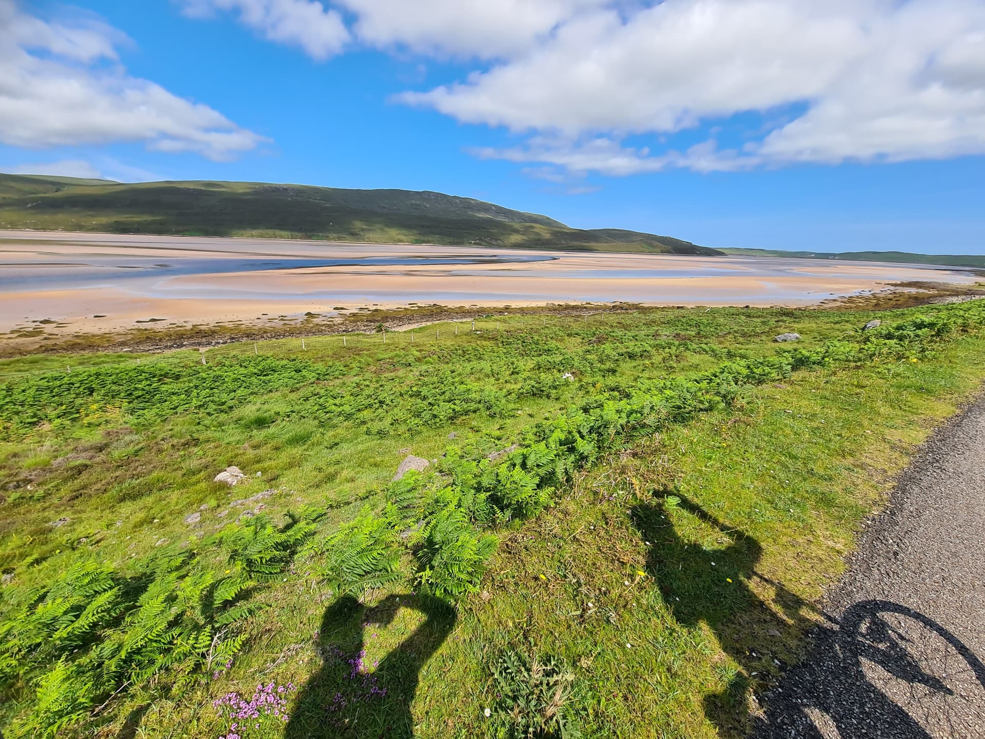 Cyclist shadow on roadside overlooking Kyle landscape with green hills and tidal flats under blue sky
